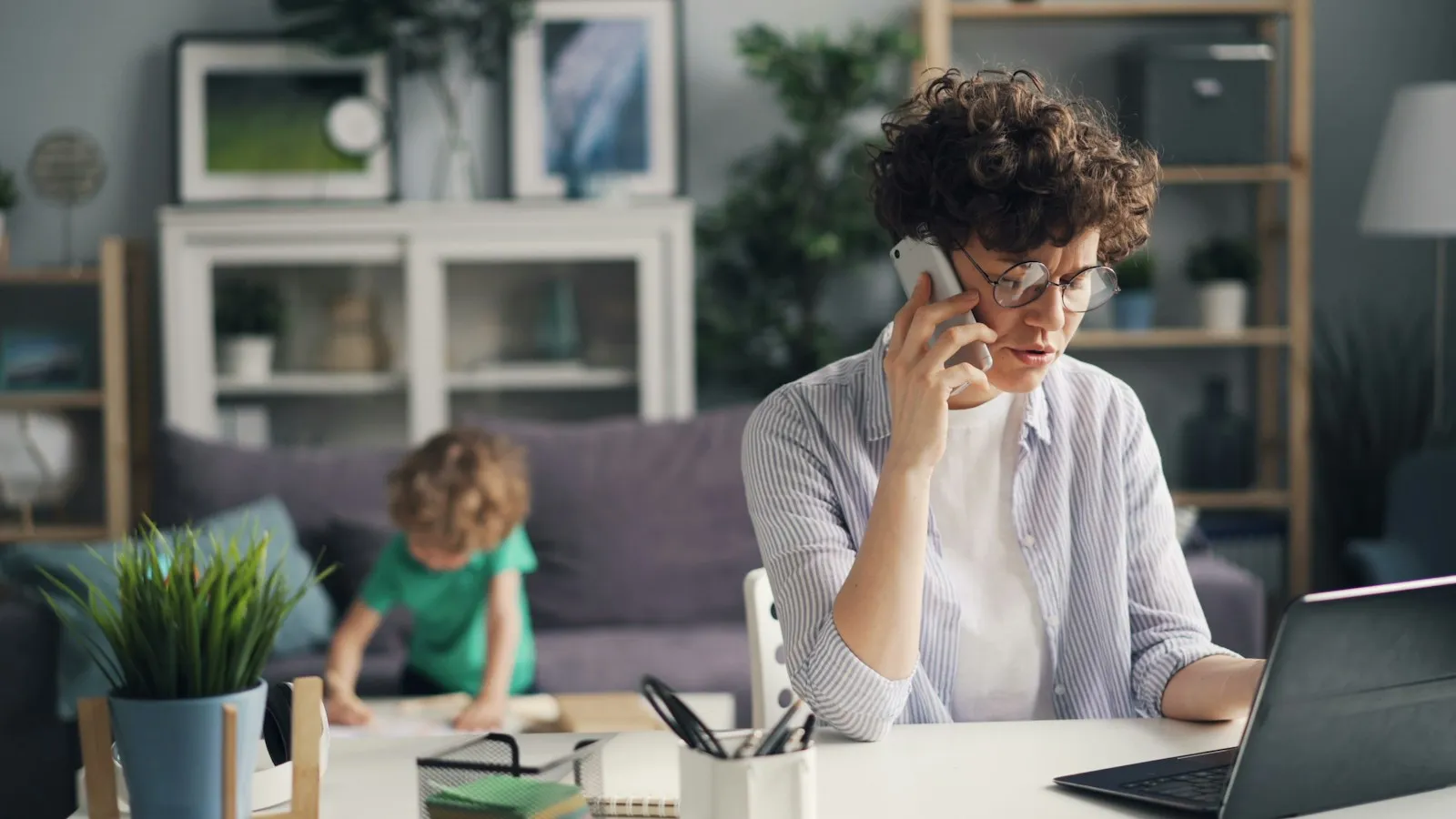 A mom working from home, on the phone at her laptop while her son plays behind her